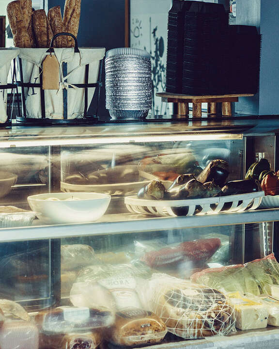 A two layer deli display case full of deli meats, cheeses, and veggies with fresh baguettes on top in a basket