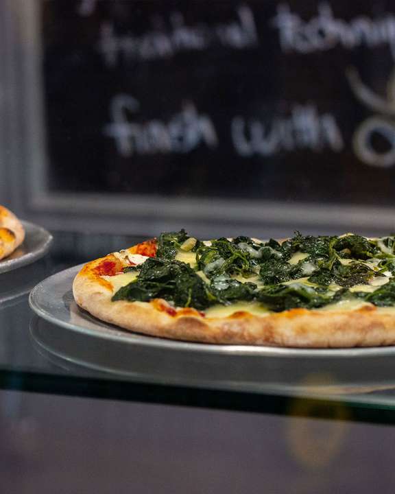 Two pizzas on metal pans, sitting on a glass shelf